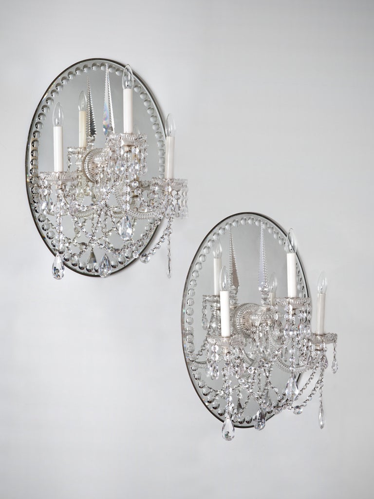 A pair of mid 19th century three arm glass wall lights. With rope twist arms with silvered nozzles and cut drip pans, each centred by a tall upward-pointing tapered notch cut prism. The whole lamp is festooned with swags of button drops and larger