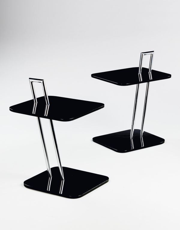 A pair of mid 20th century chrome and black lacquered wood two tiered end tables, the double column chrome support incorporates a carrying handle, each of the tiers is rectangular with rounded corners.<br />
<br />
Eileen Gray was born in 1878,