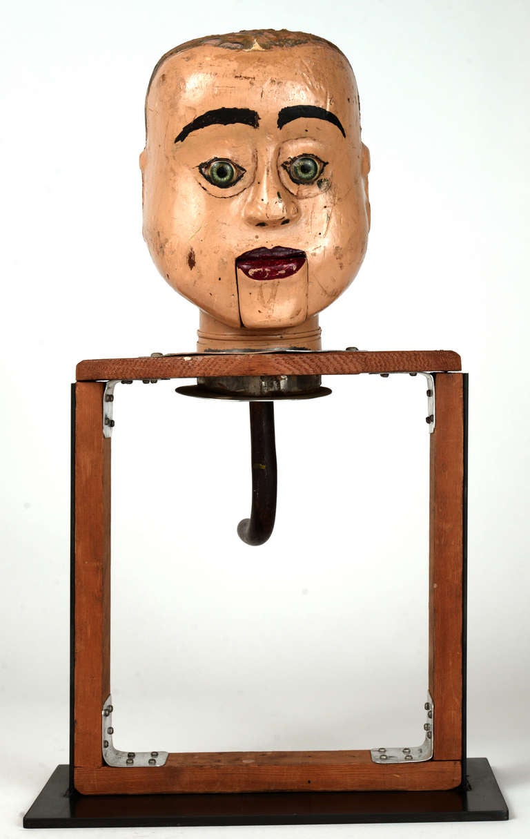 Carved wood ventriloquist's dummy head with bright blue resin eyes, heavy brows and red lips.  Mounted to a wood frame with an aluminum finger control for the mouth action and a brass handle for stabilization.  We've added a blackened steel plate