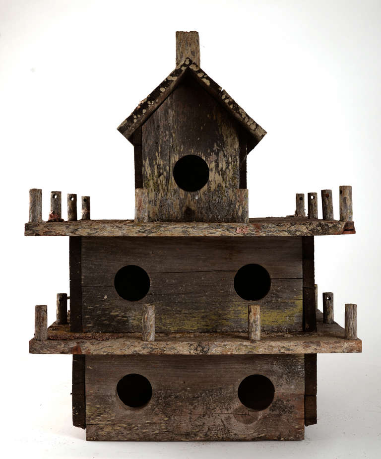 A simple, three-story bird house made of pine planks, with an  all over weathered surface including a rustic, gray color and some traces of old paint and moss.  A string of fence posts on two levels and a pitched roof with a carved chimney round out