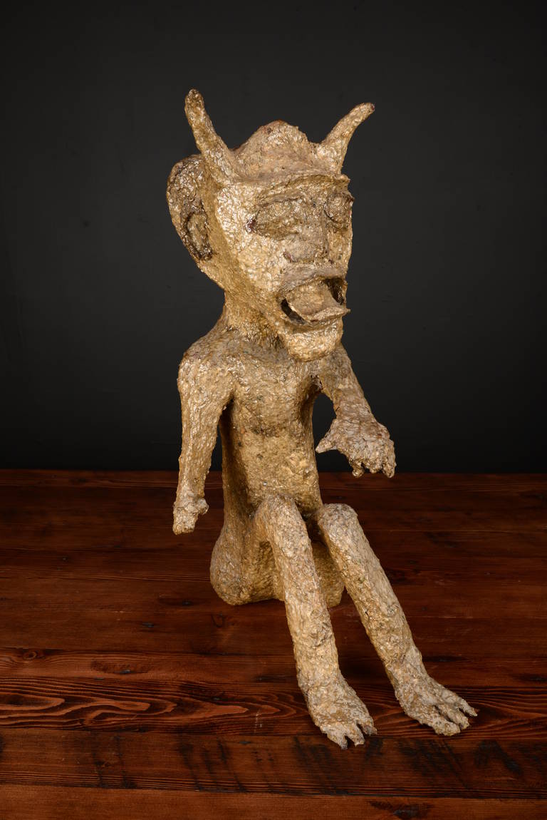 Naked seated paper mache devil with an open mouth and out stretched hand.  He's been nicely finished with a greenish-gold glaze which add to his slithery other worldly feel.