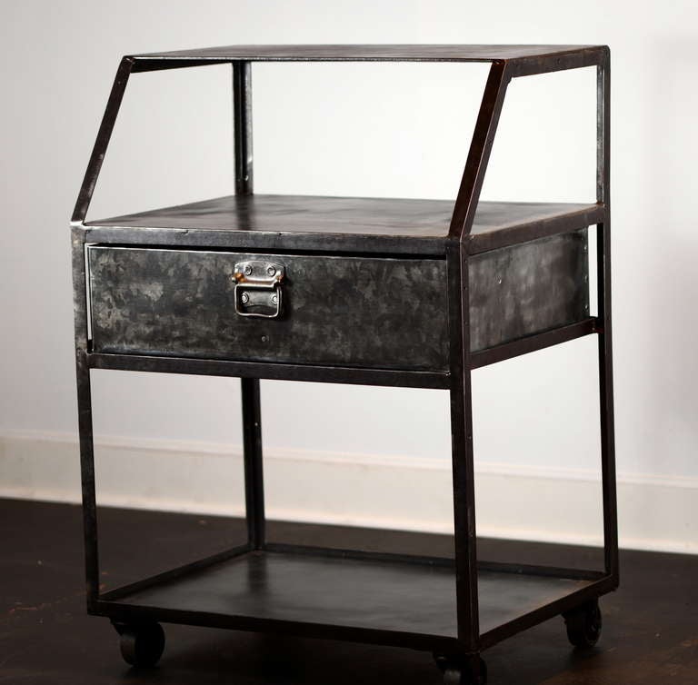 Simple steel shop desk on casters with a single full-width drawer at the center.  Slant-front rails separate a top shelf which is set back just a bit from the larger surface right over the drawer.  Lower storage below.