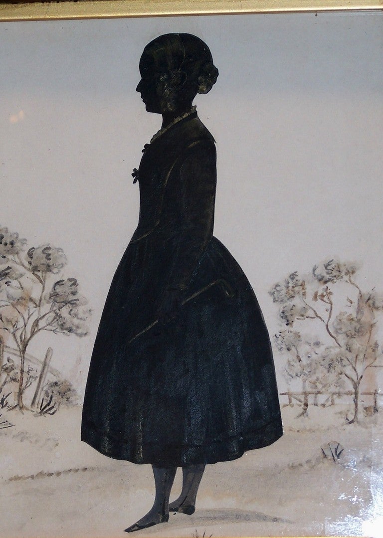A cut silhouette of a young girl with a riding crop. She wears a period dress. Her hair and clothes are detailed in Chinese white watercolor paint.
She stands in a field, the trees and wood fence in the background define the edge of the field.
The
