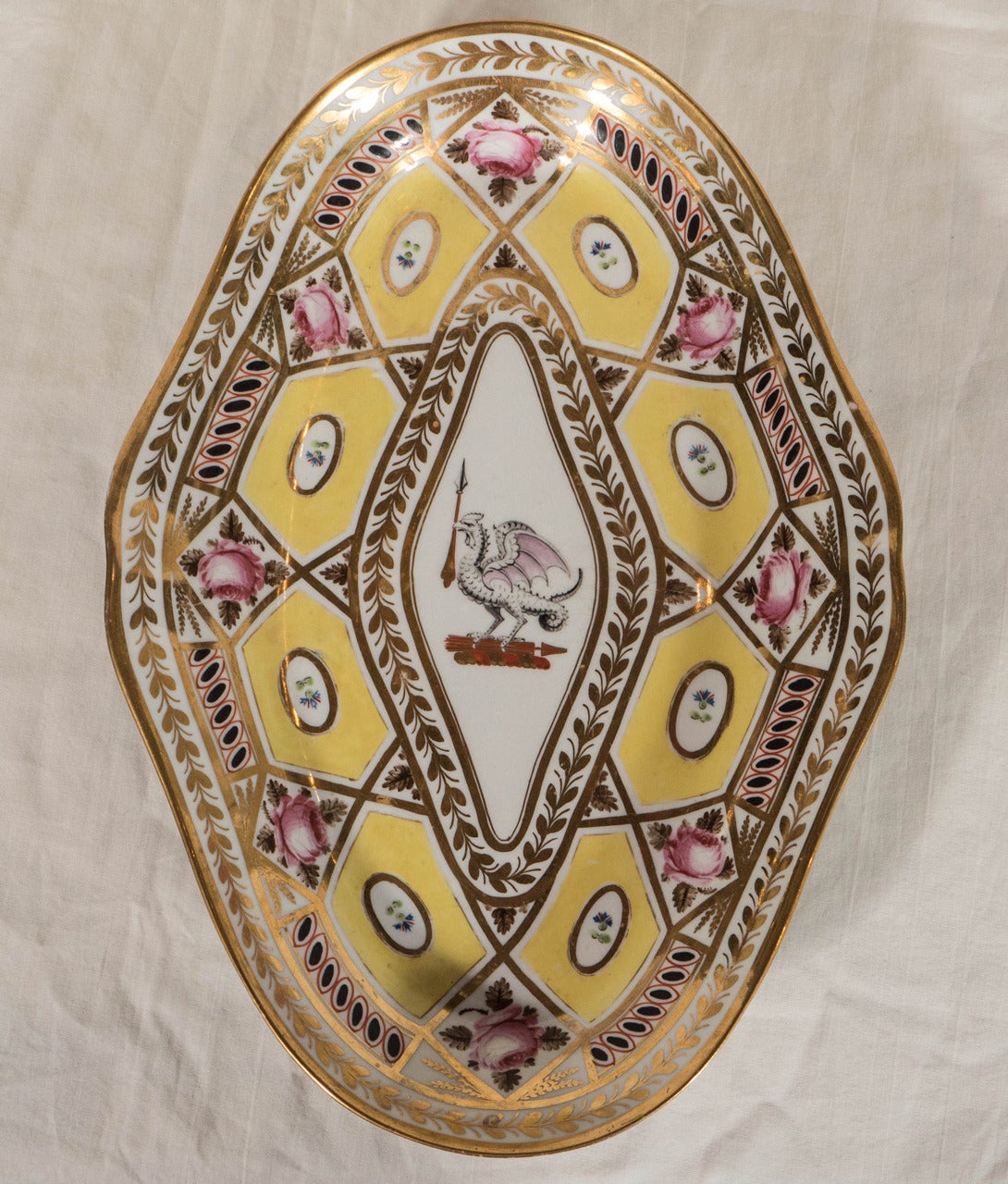 A pair of Regency period Coalport dishes in the remarkable Church Gresley pattern decorated with yellow ground hexagons, pink roses, green leaves, and exceptional gilding, all surrounding a central crest with a mythical beast. 
The crest as