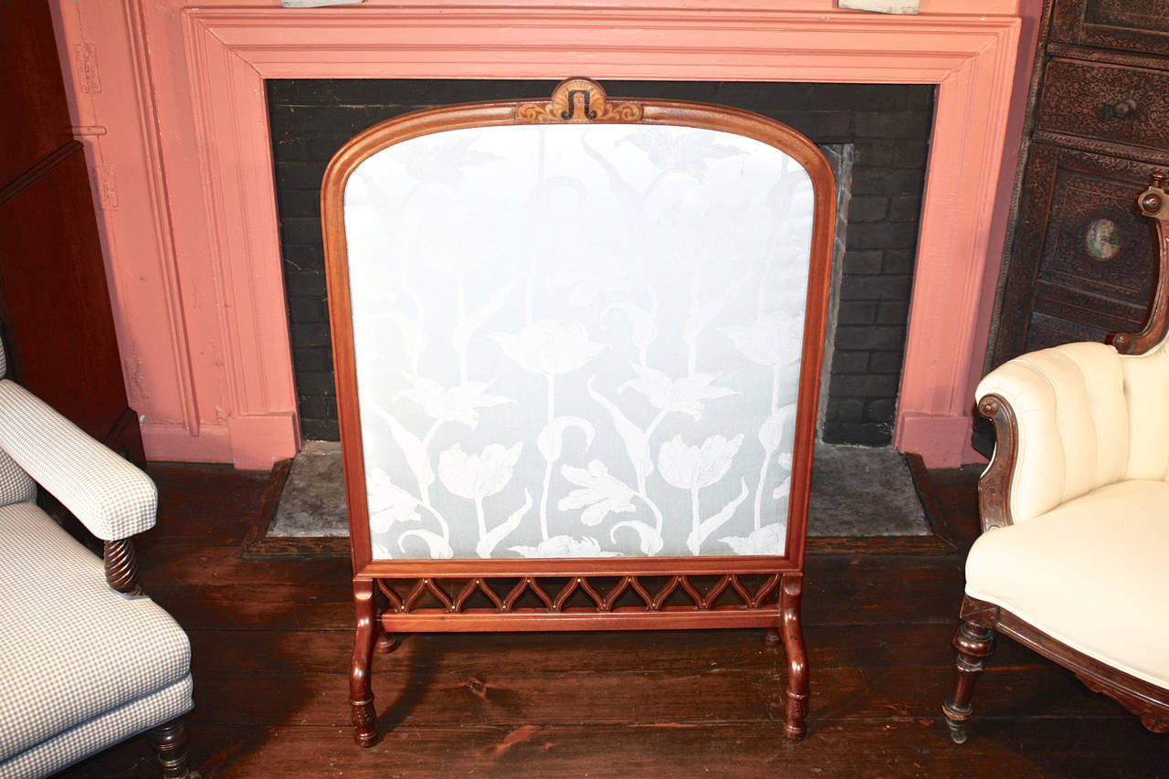 A tall silk-upholstered Art Nouveau period fireplace screen, for shielding a hearth when not in use. Its mahogany frame is intricately carved, stretchered with diagonal fretwork, and inlaid with satinwood and ebony. An inlay medallion at the centre
