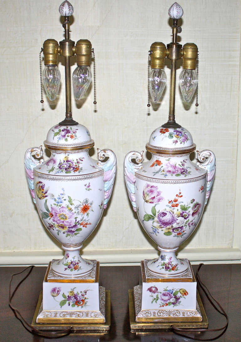 An intentional yet deliberately non-identical pair of Dresden porcelain dome-lidded urns, hand decorated in the earlier Meissen neoclassical manner; drilled and mounted on brass bases, as lamps.  The original porcelain elements, of 21 inches height,