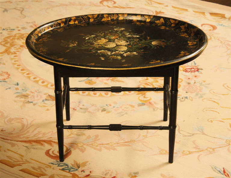An exceptionally fine painted and stenciled black lacquered oval tole tray, on a later faux bamboo turned stand.  The tray rests securely in the stand, and can be lifted out as desired.
In America, painted 'tinware' is deemed folk art; though some