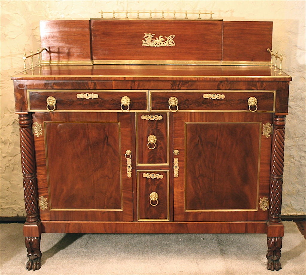 A complex, hand-carved vibrantly grained veneered mahogany, ornately brass-mounted sideboard; with gleaming back splash and gallery trim.  Attributed to the shop of Emmons & Archibald, Boston (1820-1825).  Two drawers, two cabinet doors, and two