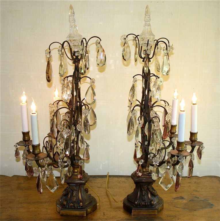 'After' a pair of Louis XVI neoclassical three candle gilt bronze girandoles, with swag decorated bases; these later French candelabra are hung with rock crystal clear, amethyst and amber pendants, suspended from clear rosettes. Topped with clear