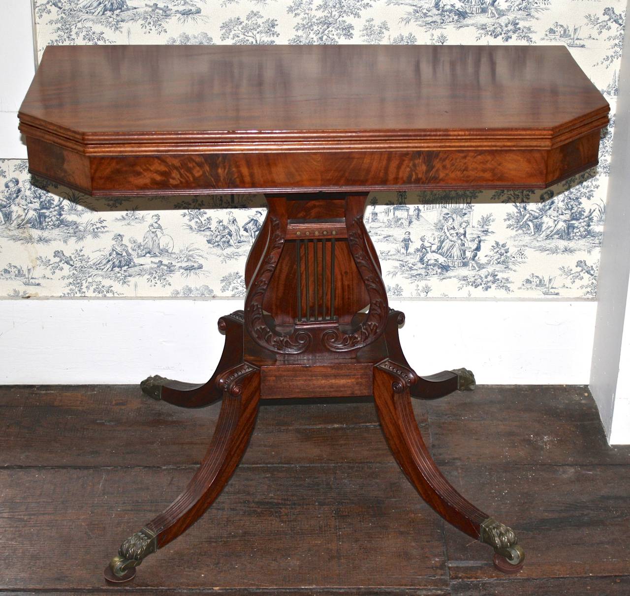A boldly figured golden mahogany fold-over games table, on a foliate carved and brass strung lyre-form base. Reeded legs with trimmed knees and original brass pawed casters. The fold-over top has a moulded edge and diagonal hypotenuse-cut corners;