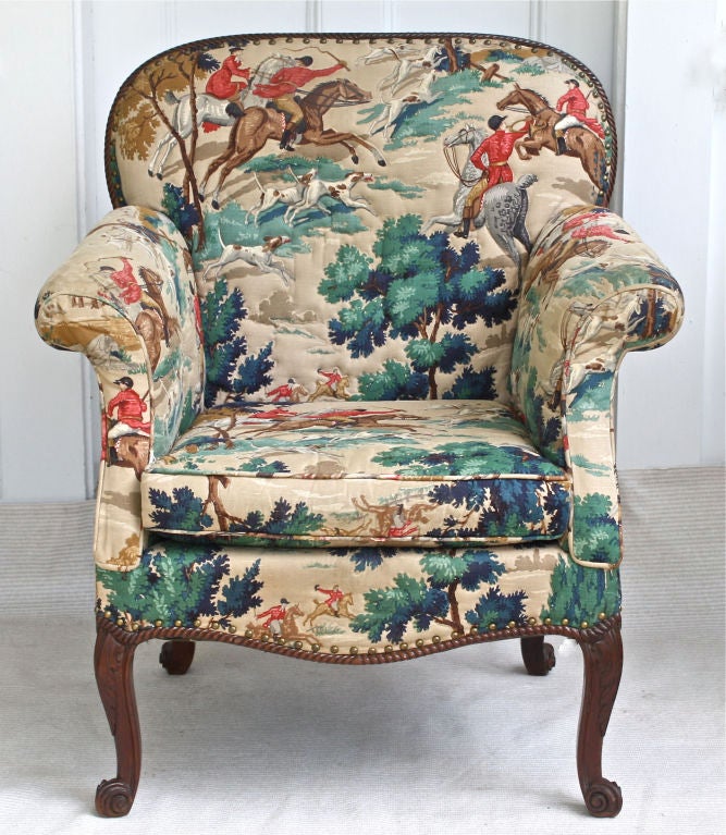 An upholstered library armchair with gadroon carved balloon-back and apron framing; on front and back French scroll carved feet.  Examination of innards reveal that it is an antique Georgian Revival chair, likely of the mid to late nineteenth