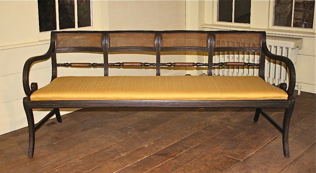 A four-chairback Federal period long settee, in its original black-green paint, parcel-gilt trim, and caning; with a newly upholstered yellow gold herringbone stripe slip seat, conforming to its frame contour. This form of Klismos chair and settee