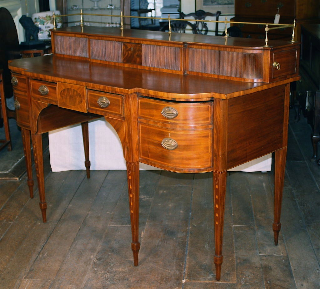 A Hepplewhite manner sideboard of richly grained honey colored mahogany, with complex string and bellflower satinwood inlays, and crotch mahogany contrasting ovoid and rectangular plaques.  Its top is surmounted by a tambour-faced 