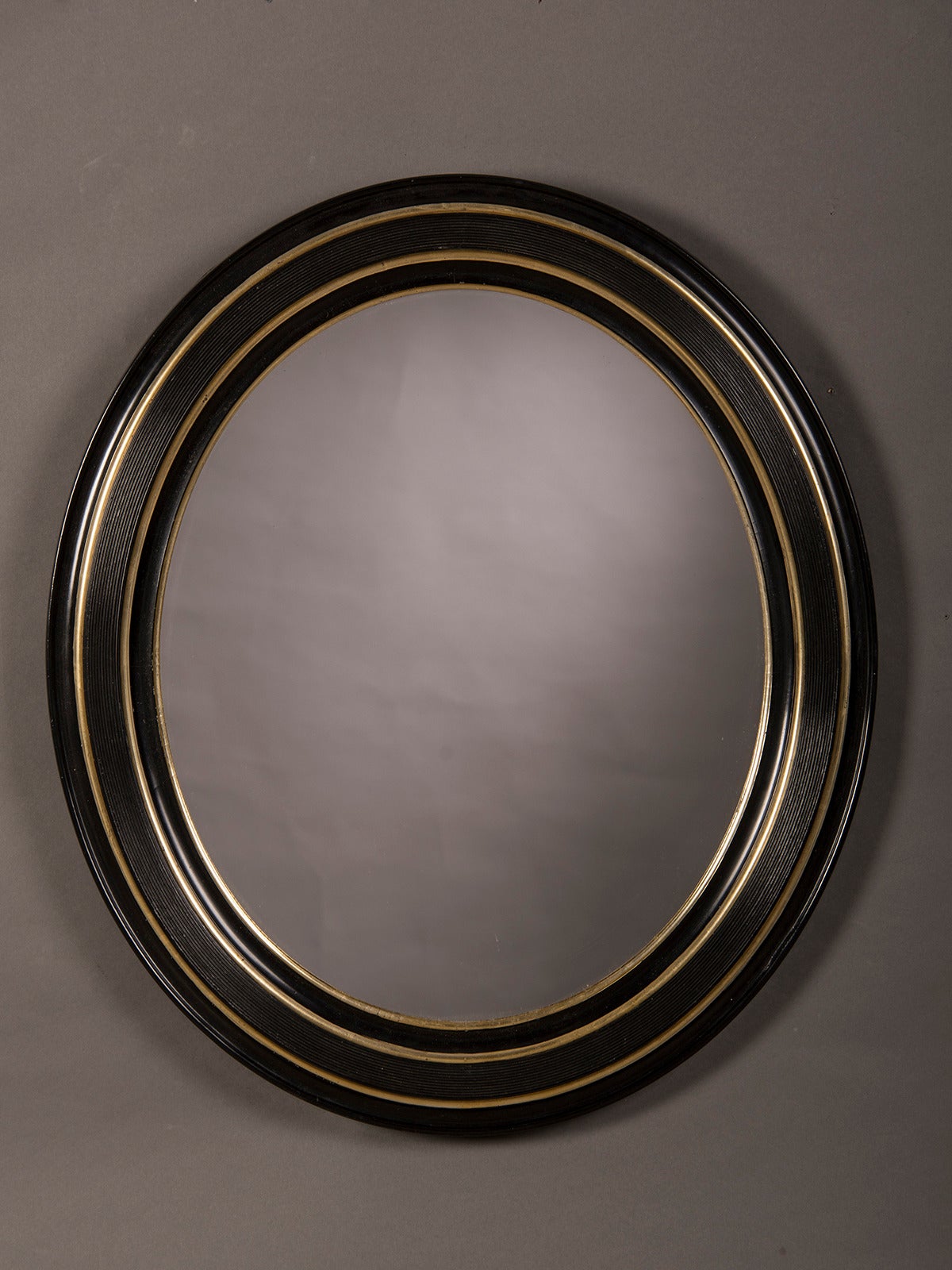 A handsome oval wooden frame lacquered in black with gold leaf highlights enclosing the original mirror glass from England c. 1890. Please notice the three alternating bands of black and gold with each having a different scale and reflective value.