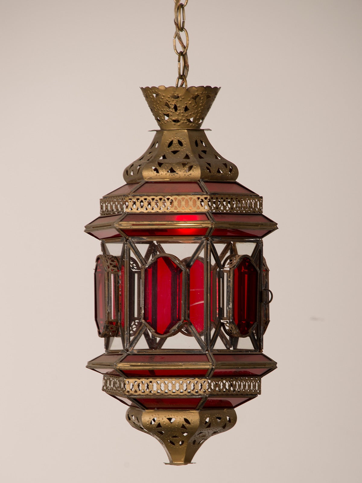 A vintage Moroccan lantern circa 1940 enclosing ruby and clear glass within a framework of pierced and filigreed brass. This lantern has a hexagonal shape and the interplay between the coloured glass and the textured brass creates a romantic aura of