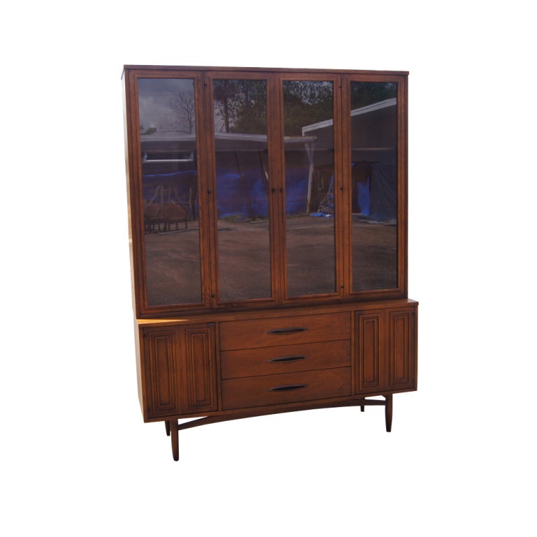 A mid century modern walnut Sculptra series china cabinet made by Broyhill.  The lower section with three drawers and two doors concealing shelved storage. The upper section with four glazed panels and shelves.