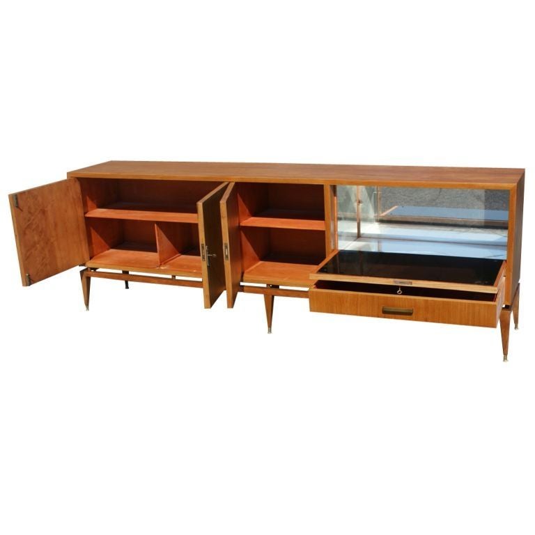 A mid century modern buffet or sideboard made of walnut.  Three doors concealing adjustable shelving, a fitted drawer for flatware, and a drop front, lockable bar with mirrored interior.  Brass hardware and sabots. Small blemish on top.
