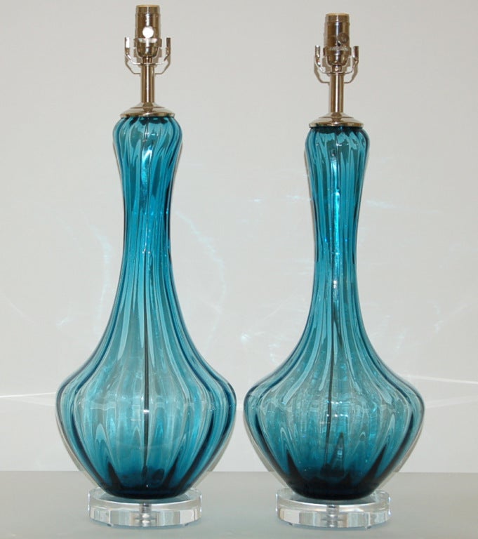 A spectacular pair of TEAL BLUE Murano lamps with vertical optics. The color of the glass is what really makes these lamps special!   We show them on chunky Lucite paired with nickel hardware - simply elegant. 

The lamps stand 27 inches from