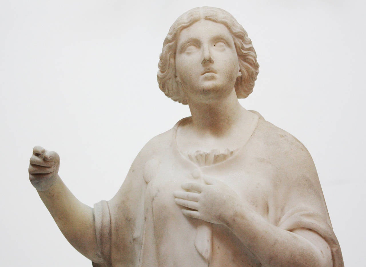 a white marble sculpture of a young woman with a short cape buttoned over a loose classical chiton, she wears a clam shell on her right shoulder, her hand is outstretched, the other over her heart, signed TC (monogram / cypher) on back base

cult