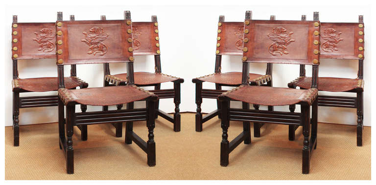 Set of six Spanish Renaissance Revival style chairs with rust leather backs and seats, each seat back is embossed with a rampant lion with wings and each seat a woven pattern, large round brass nail heads decorate the edges.