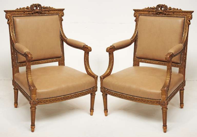a pair of elaborately carved giltwood framed French Louis XVI style arm chairs, floral wreaths and swags on crest rails, new buff leather upholstery