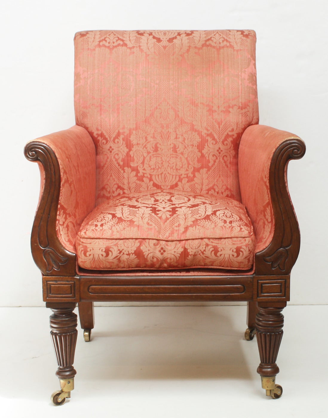 An upholstered English Regency mahogany library chair with wood arm fronts with bellflower design. Reeded front legs with casters.