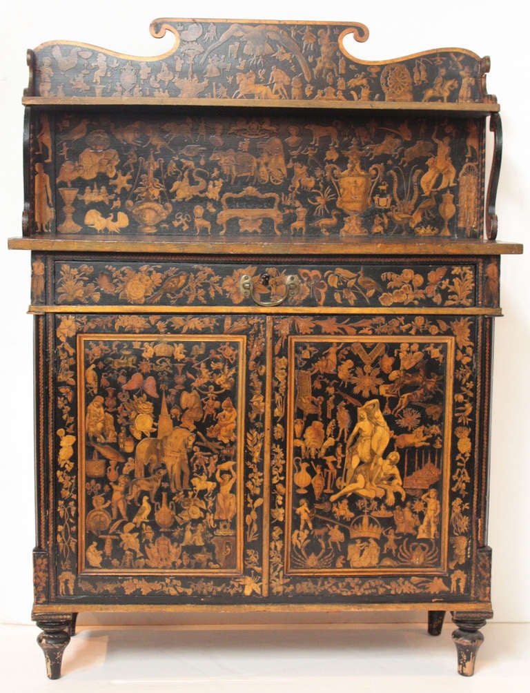 a small chiffonier with shaped top, shelf supported with scroll forms, large single drawer over cabinet doors, painted black then decorated overall with cut out paper pictures of birds, butterflies, flowers, people and animals, the whole varnished