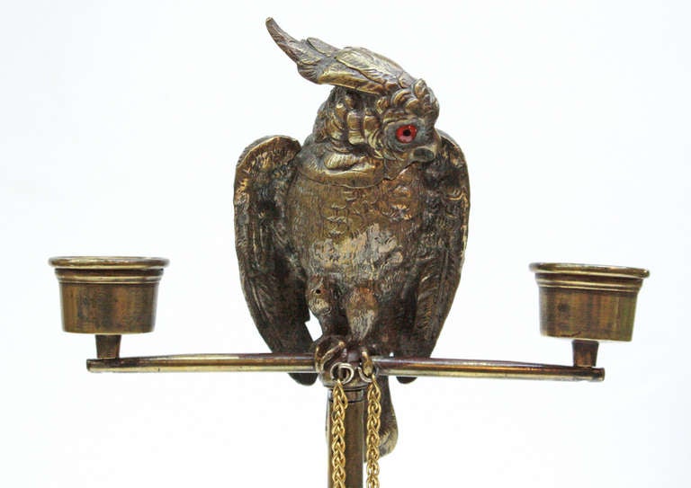 a sculpture in the form of a parrot chained to a perch whose head lifts to provide a match holder, two cups, one at each end of the perch could be used for candles, the bird has glass eyes