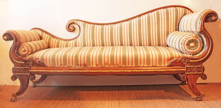 a period Regency recamier with carved walnut frame, curving frame is embellished with gilt metal accents, terminating in anthemion mounts above feet with castors, the whole upholstered in a very tasteful stripe in creme and celery with rust (some