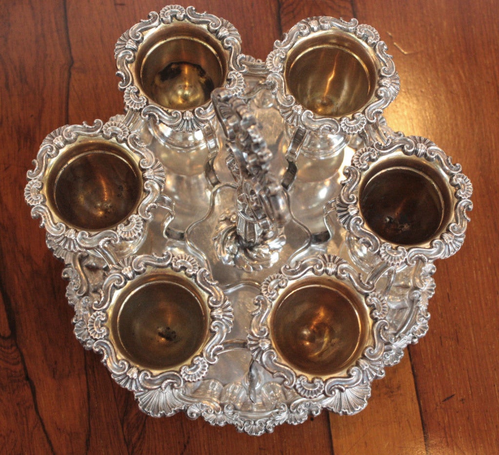 a Sheffield plate egg cruet with six cups with elaborately chased edges. Interconnected holders stand on heavily chased tray on feet. Center handle