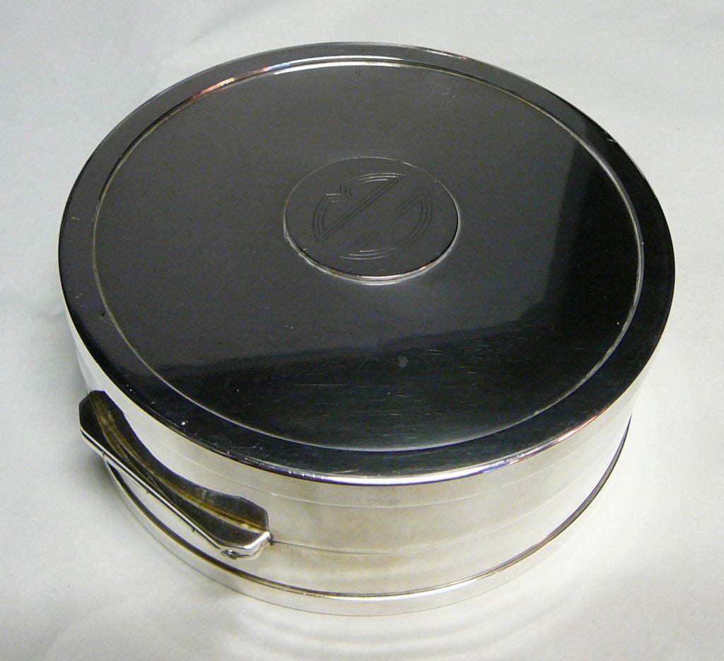 A stylish silver-plated circular trinket box by Yves St. Laurent, signed on the base, the drum body hinged at the centre, the lid with a circular cartouche engraved in Deco script with the initials ‘C B’.