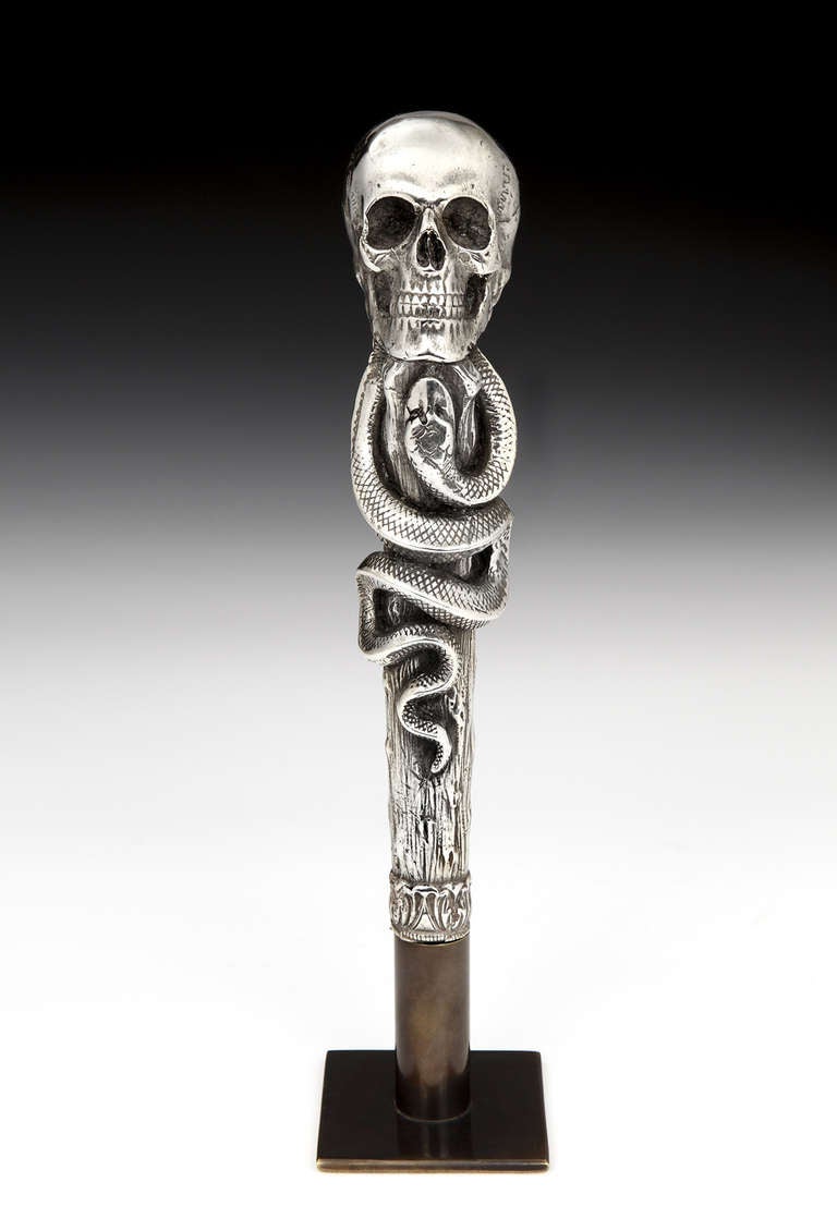 A superbly modelled and highly decorative silvered bronze cane handle in the form of a skull and serpent, the handle set on a bespoke stand to enable it to be displayed ‘off’ the cane'. German, circa 1930s. Height on stand: 9 inches (23 cms).