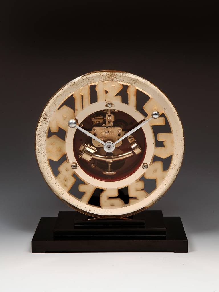 A handsome glass Art Deco ‘Skeleton’ mantel clock, unusually with the numerals formed from frosted glass and contrasted against the pale pink tinted mirror face rather than in applied chrome against clear glass. With silver-plated ‘spoon’ finial