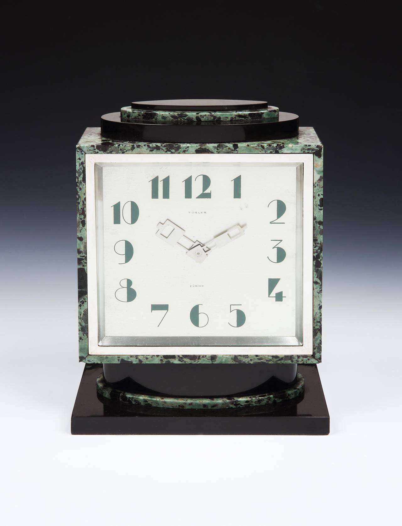 A strikingly modern Art Deco mantel clock, the main body in verde antico marble, with architectural, stepped detailing in polished black marble. The brushed, silvered-bronze clock face with modernist numerals, accented by hands of a pierced design.