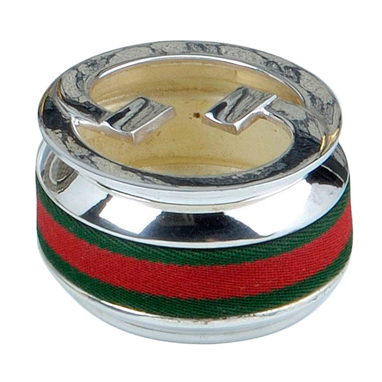 A stylish circular silver plated ashtray by Gucci, with a fabric band around the body in the signature Gucci colours of green and red, the removable lid with two interlocking ‘G’s, integrating two cigarette rests. 

Marked GUCCI MADE IN ITALY to