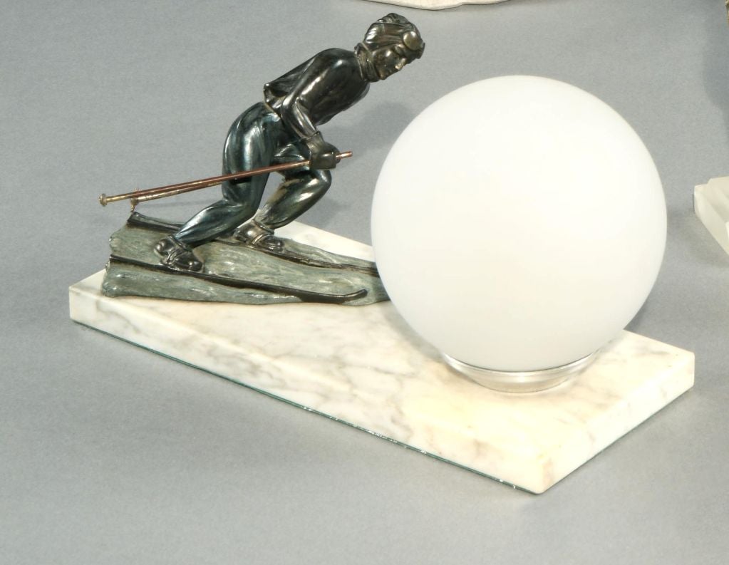 A stylish Art Deco table lamp featuring a cross-country skier on a bronze socle, the ‘terrain’ in white marble and a large white glass ‘snowball’ as the lamp.