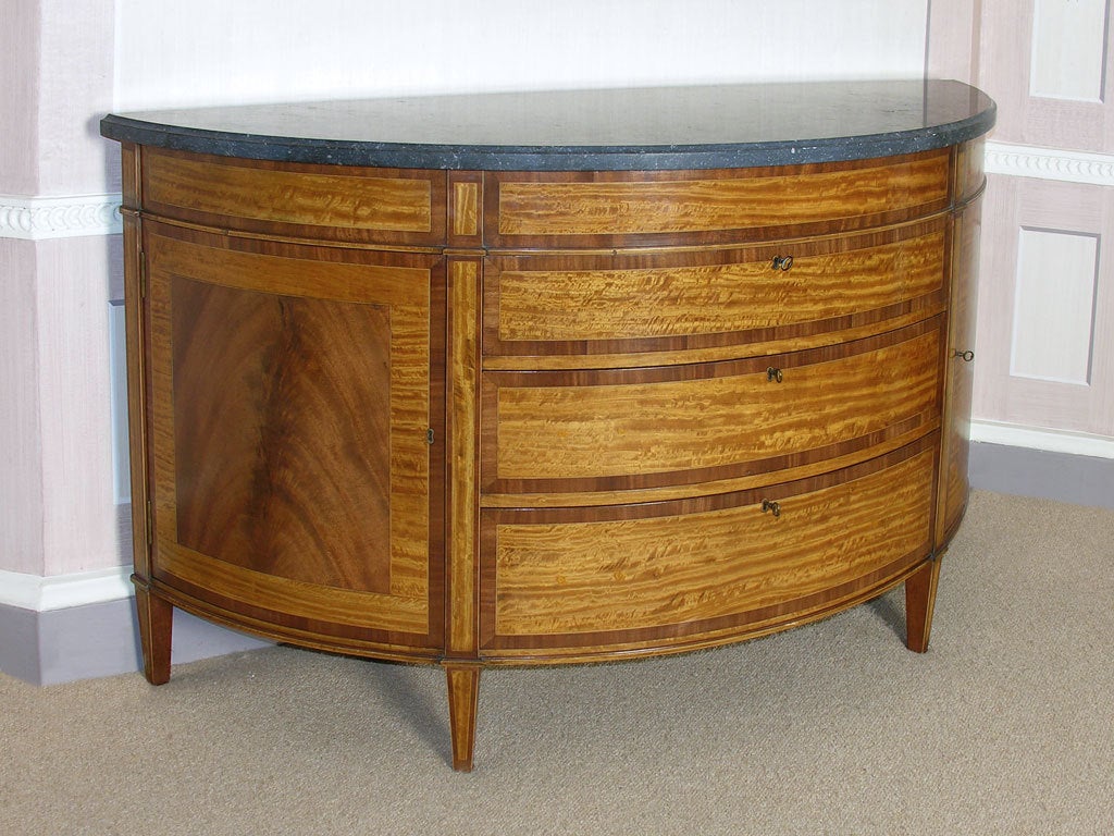 A 1920s satinwood and mahogany demi-lune commode with three crossbanded drawers and a later stone top.