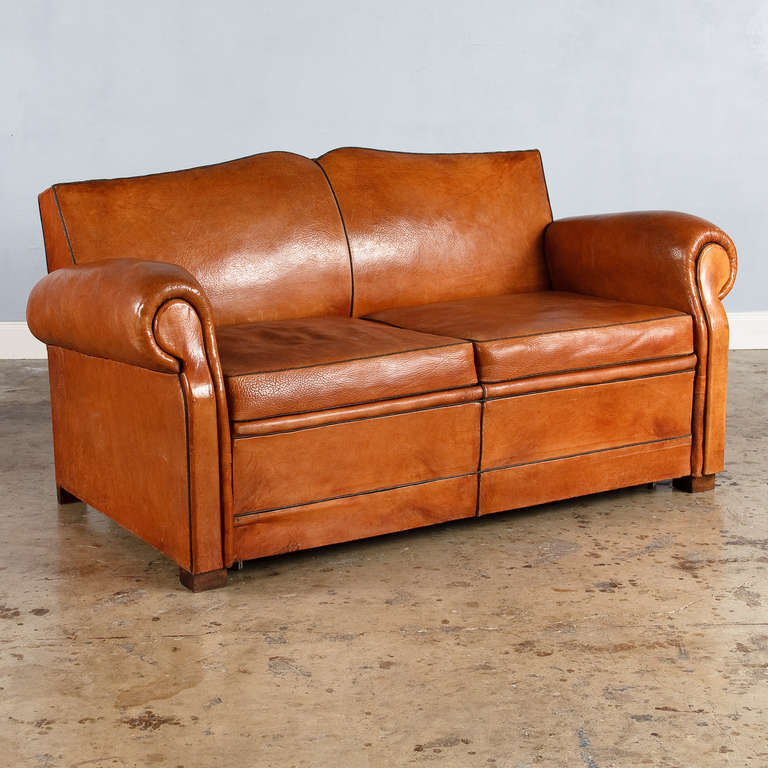 A beautiful 1930s leather club sofa, Mustache style, with a gorgeous old patina. The sofa has two cushions and rests on wooden feet. The back is upholstered in brown fabric with antique nails. This is a sofa bed that will open up with a metal frame