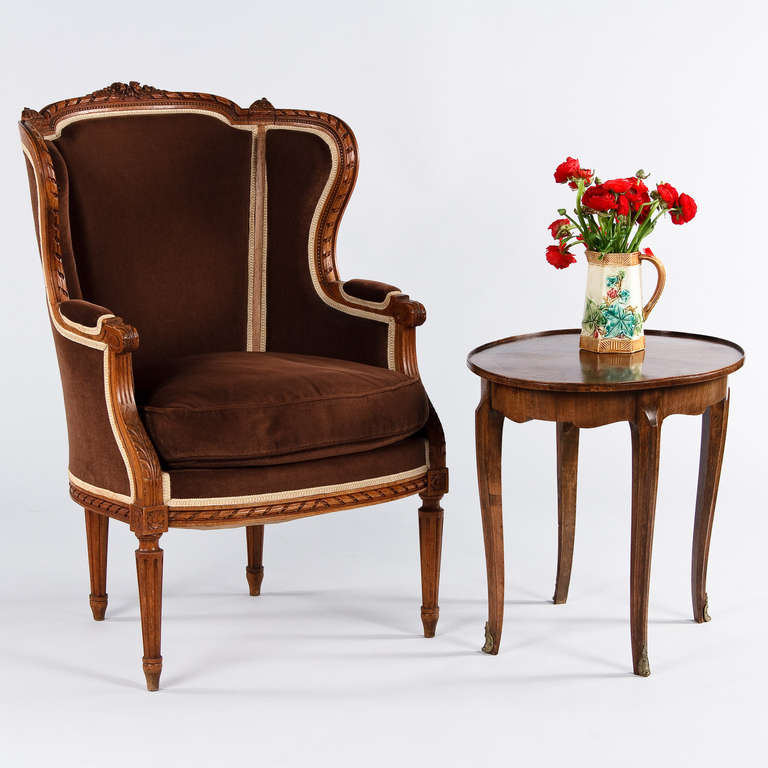 A Louis XVI Style Bergere Armchair in carved beechwood with acanthus leaves, rosettes, beading and fretting motifs. The crests feature carved flowers and the legs are fluted. The Armchair has been upholstered in chocolate brown fabric. Floor to