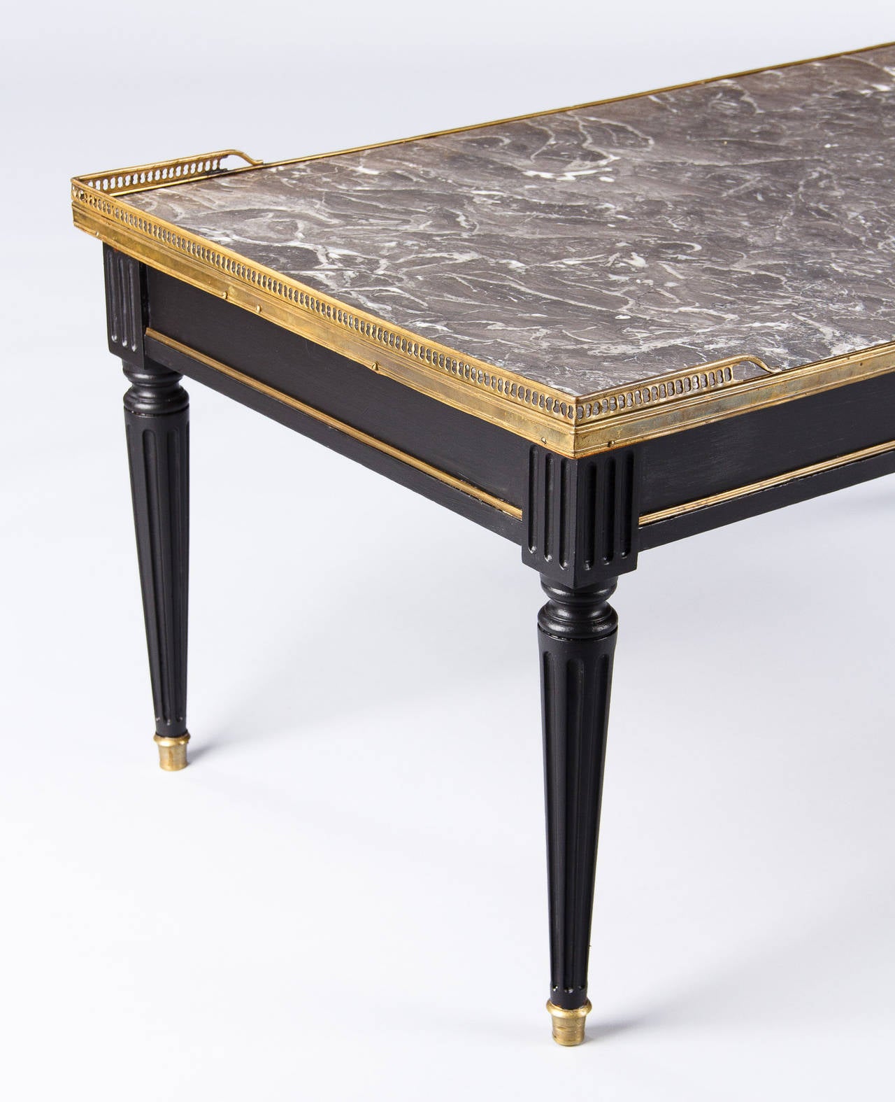 An elegant coffee or cocktail table in the Louis XVI style painted black on mahogany with a brass galleried top and trim around the apron. Also featuring brass sabots at the base of the fluted legs and a veined grey marble tabletop.