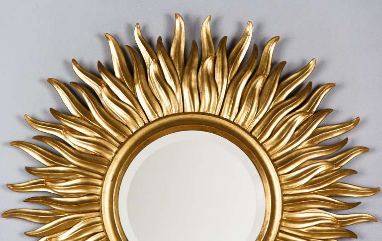 An outstanding French Mid-Century Gilt Wood Sunburst Mirror. From the south of France, this hand-carved wooden sunburst mirror features a round beveled center glass and is framed with a simple sculpted frame with three rows of flamed sun rays with