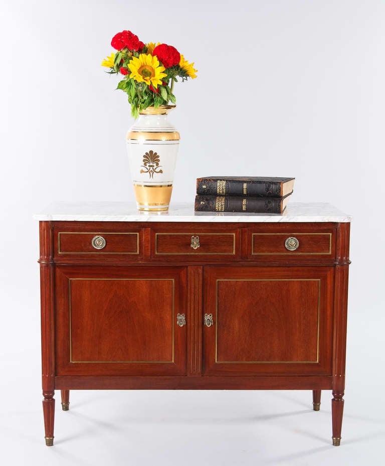 A wonderful Sideboard in the Louis XVI Style made of lacquered red mahogany with a white Italian Carrara Marble top with rounded edges. The Sideboard has perfect proportions and features two doors and three drawers. The door panels have elegant