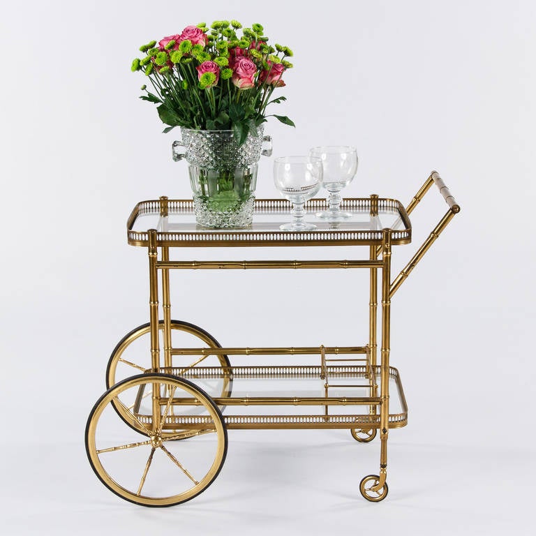 A midcentury brass bar cart in faux bamboo style from Maison Bagues. The cart features two clear glass tops with brass galleried edges and a three bottle holder. The shaped wheels are brass with hard rubber tires.
