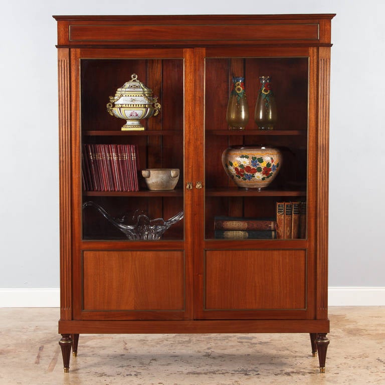 A Louis XVI style two-door bookcase in blond mahogany with brass trim. The bookcase has simple, very clean and elegant lines, unusually wide for a two-door piece. The tapered feet are fluted and finished with brass sabots. The inside has three