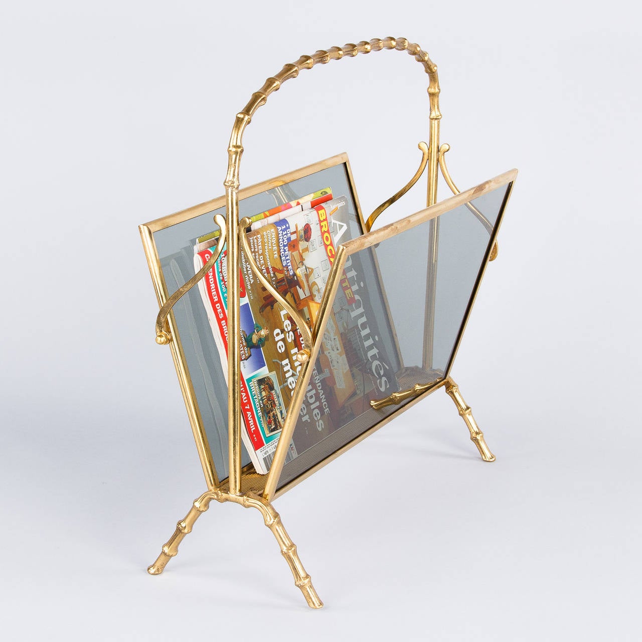 A Mid-Century magazine holder from Maison Baguès made of brass with smoked glass panels. The brass frame features scrolls motifs and bamboo style handle and feet.