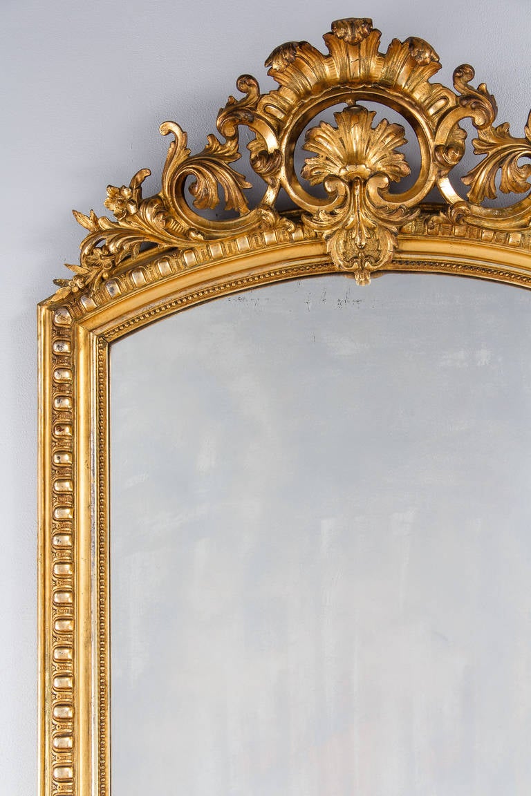 A fabulous French Louis XV-Louis XVI Transition period mirror in gold leaf with its original glass. The gilded frame has gadrooned moldings and a beaded trim around the glass. The arched top crest has open work intricate carvings in shells,