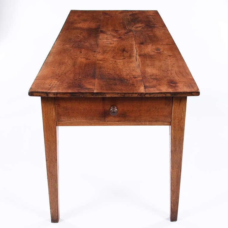 A warm and rustic French Country Farm Table from the Rhone Region made of pine wood and oak tapered legs. The table features a drawer at one end and another drawer on the side.  Both drawers have hand carved wooden pulls. Perfect for a dining room,