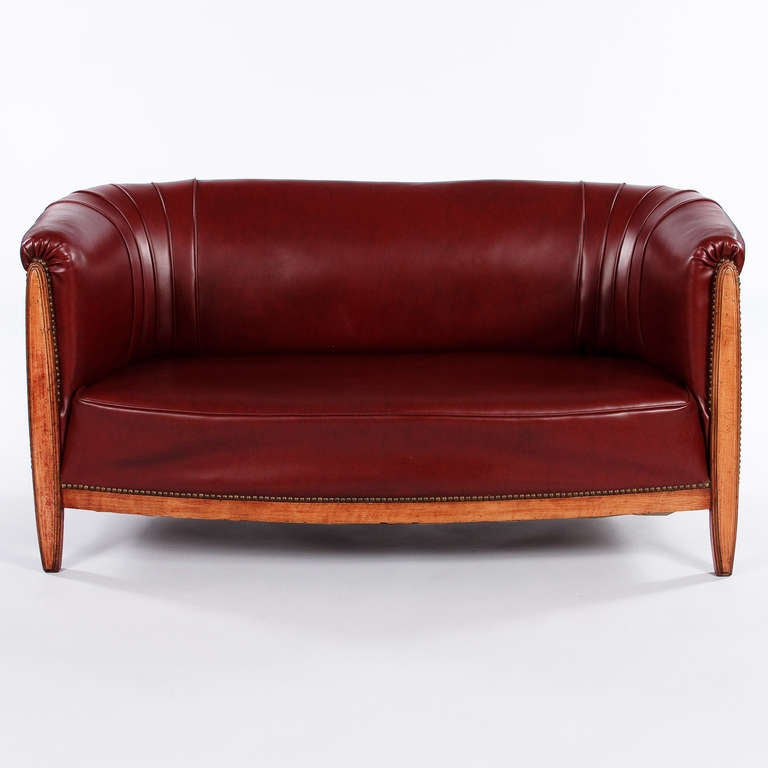 A small sofa from the French Art Deco Period with a curved back upholstered in a rich burgundy red vinyl with antique nails. The frame is cherry wood. A perfect fit for a child's room, at the foot of a bed, or as a window seat!