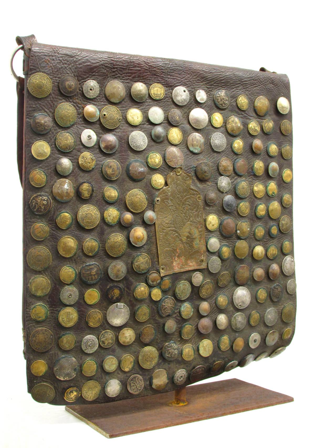 Large leather bag with flap decorated with domed and riveted coins surround a brass hand etched mihrab shaped form the coins vary in age from late 19th century through 1970s displayed on custom-made stand.