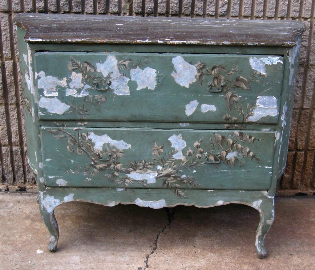 Old crusty antique two-drawer commode with curved sides with multiple layers of worn and weathered paint.